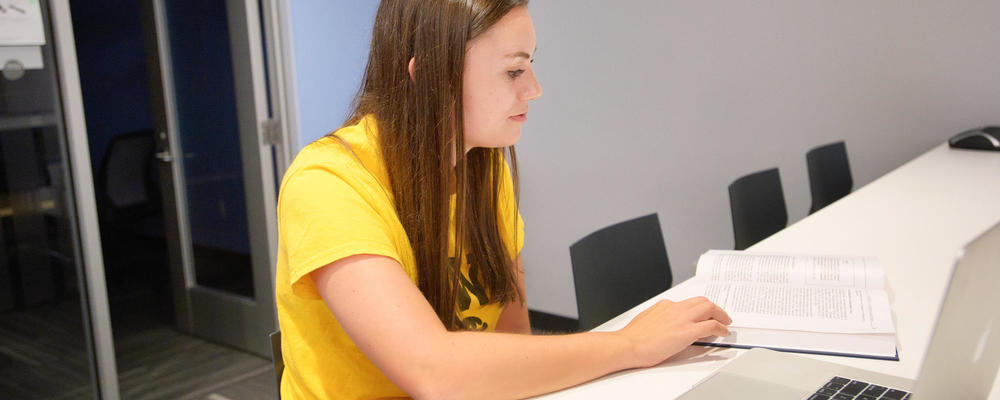 Student wearing a yellow shirt looking down at an open text book with an open laptop next to the book