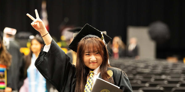 a student in cap and gown throws up a peace sign and smiles