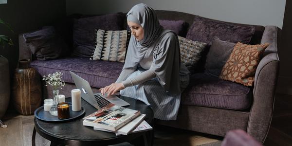 person wearing hijab sits on a purple couch and uses a laptop