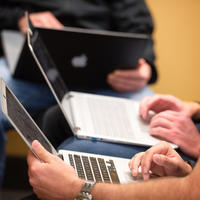 zoomed in on three sets of hands holding and typing on laptops. The focal point is of a man's hands wearing a silver watch holding a macbook pro